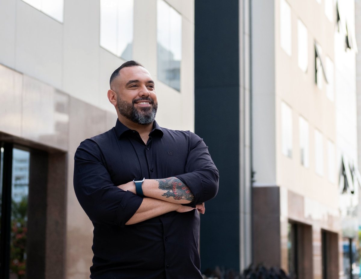 JC sorrindo, de braços cruzados, em frente a um prédio comercial