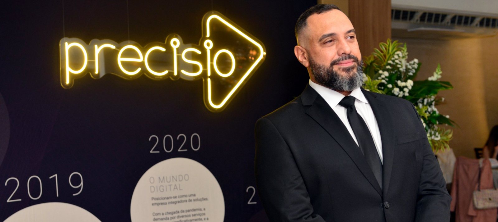 JC em evento da Precisio, diante do painel com a linha do tempo da empresa